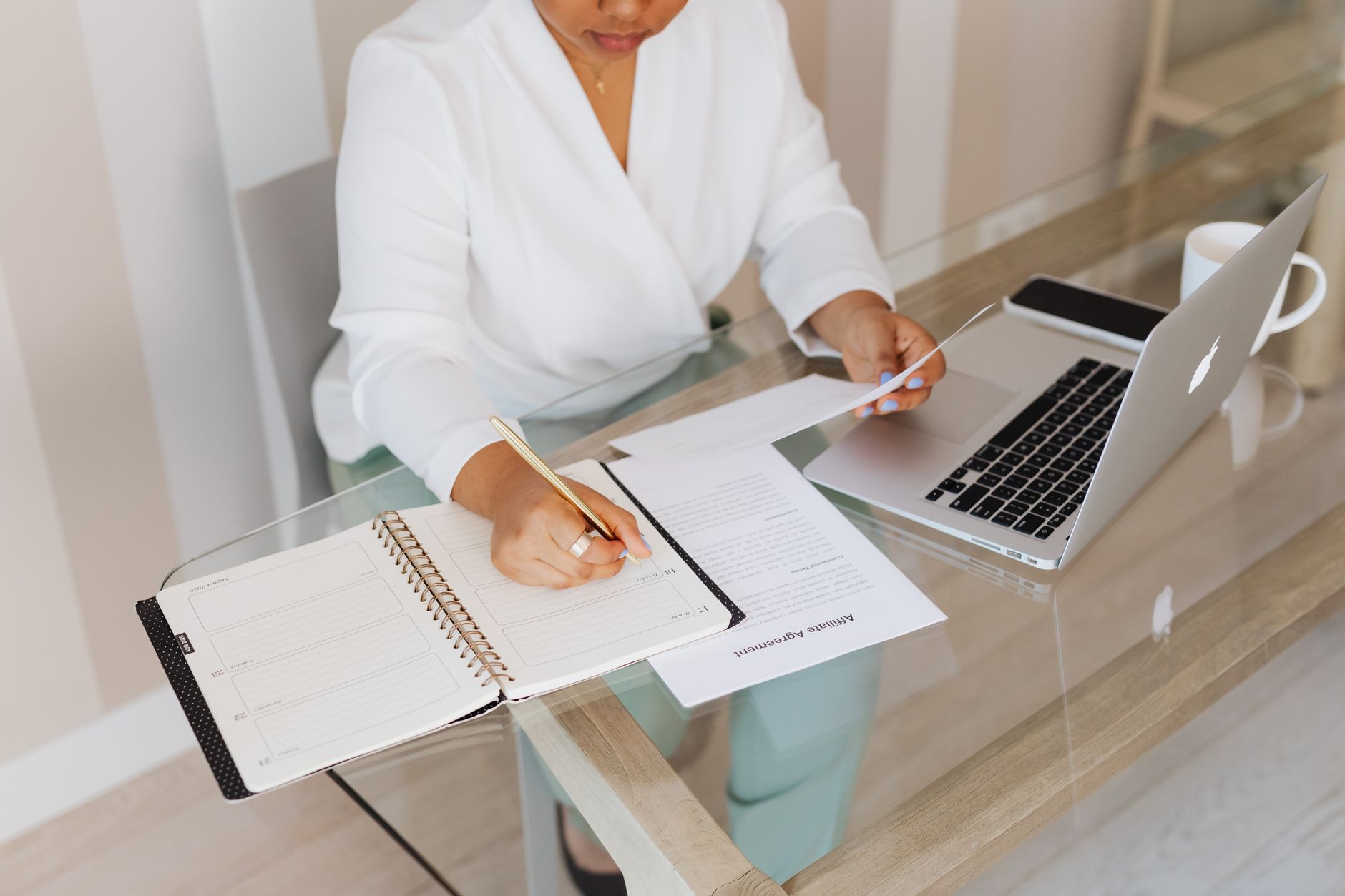 Femme prenant des notes tout en travaillant avec un ordinateur portable et des documents à un bureau en verre à l'intérieur.