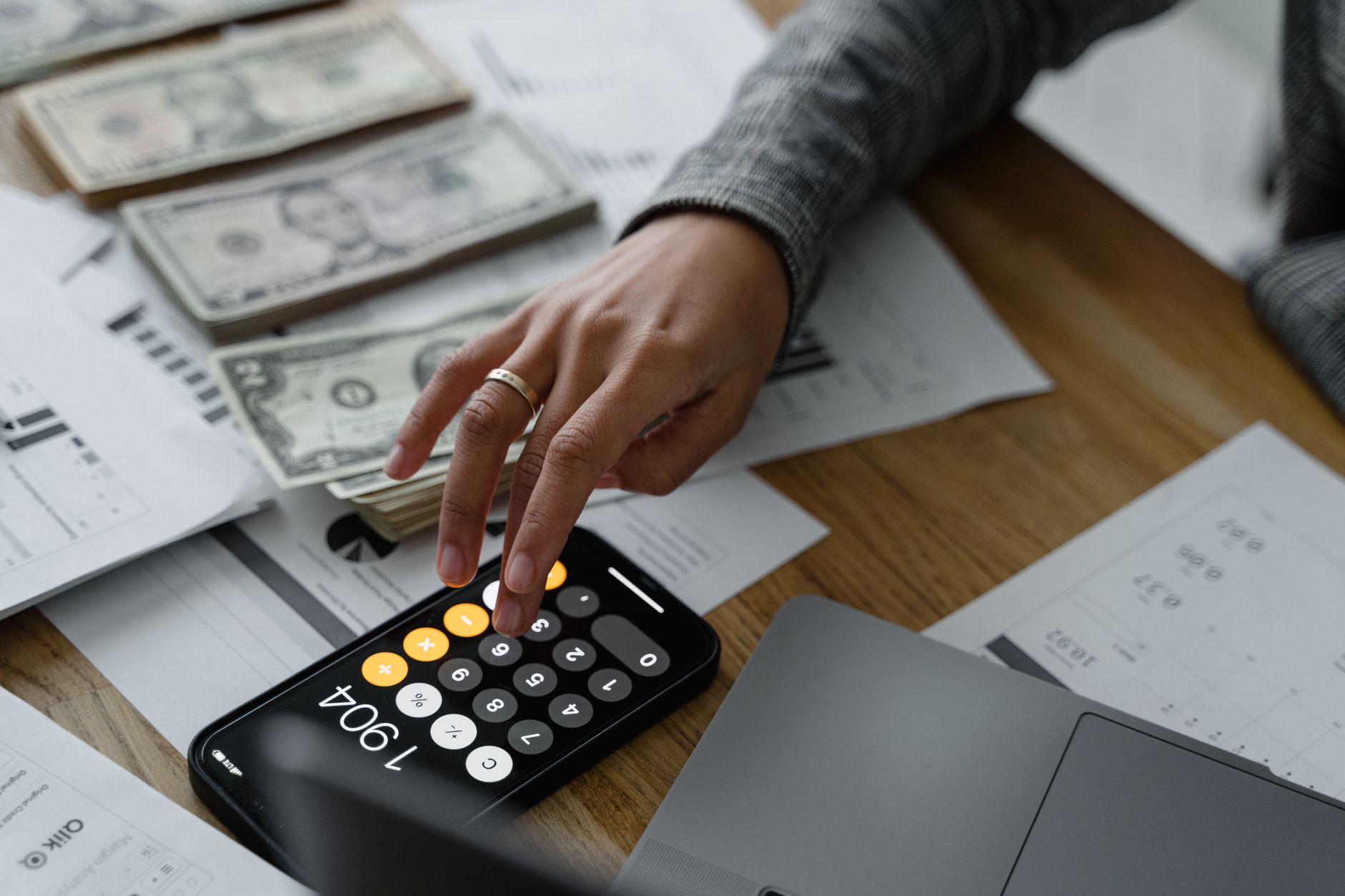 Gros plan d'une personne utilisant une calculatrice sur smartphone au milieu d'argent et de documents financiers sur une table en bois.