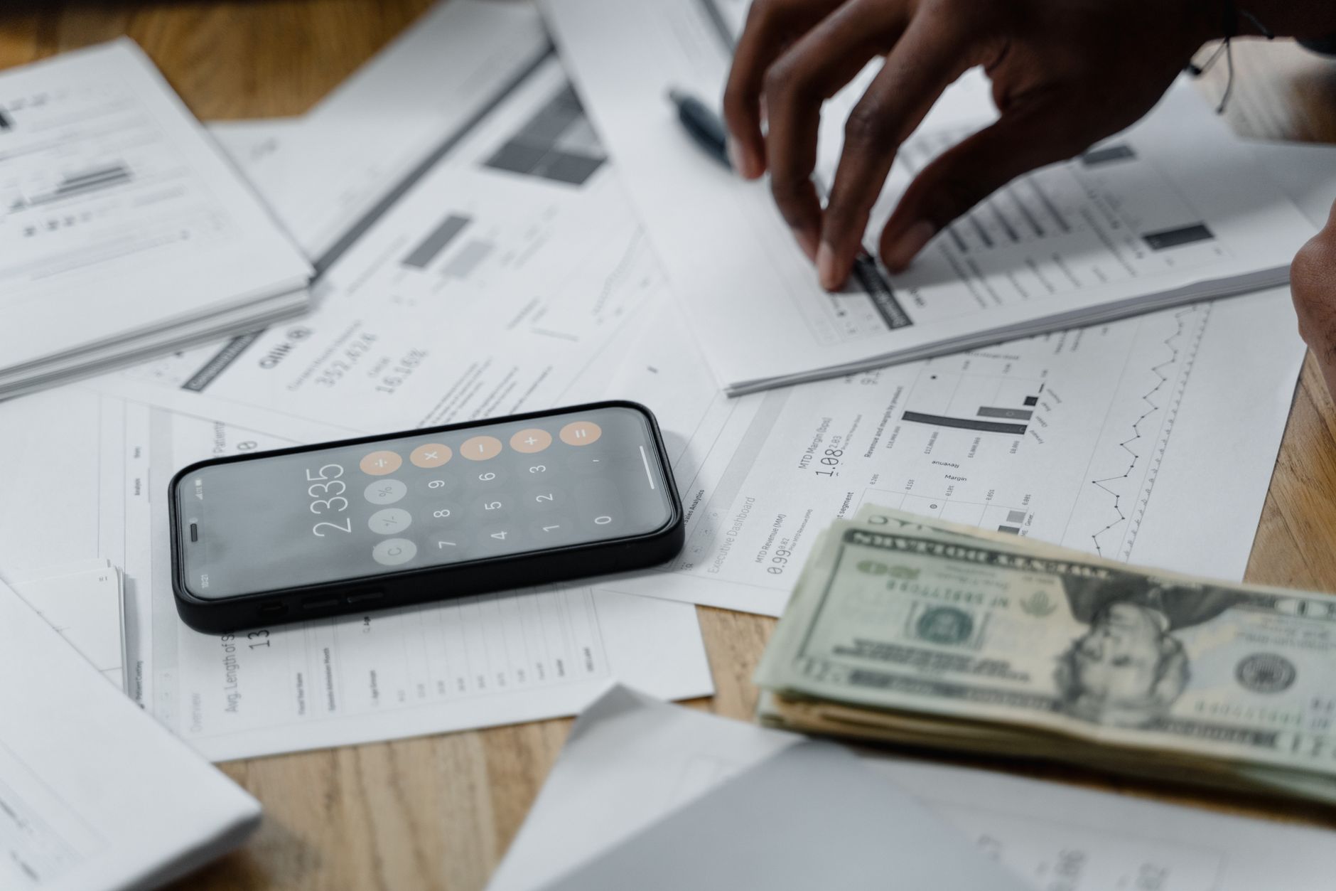 Documents financiers, liasse de billets et smartphone avec calculatrice posés sur une table en bois.