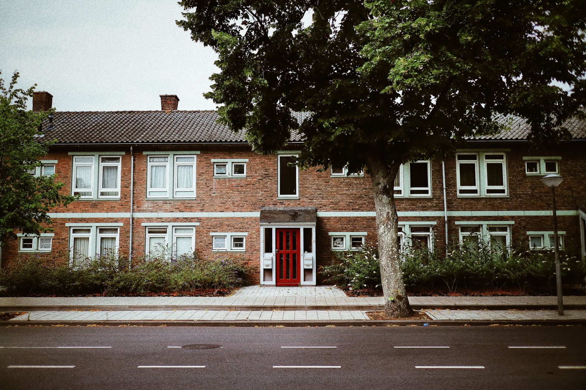 Façade en brique rouge d’un immeuble résidentiel à deux niveaux, avec porte rouge, grand arbre sur le trottoir et route vide au premier plan.
