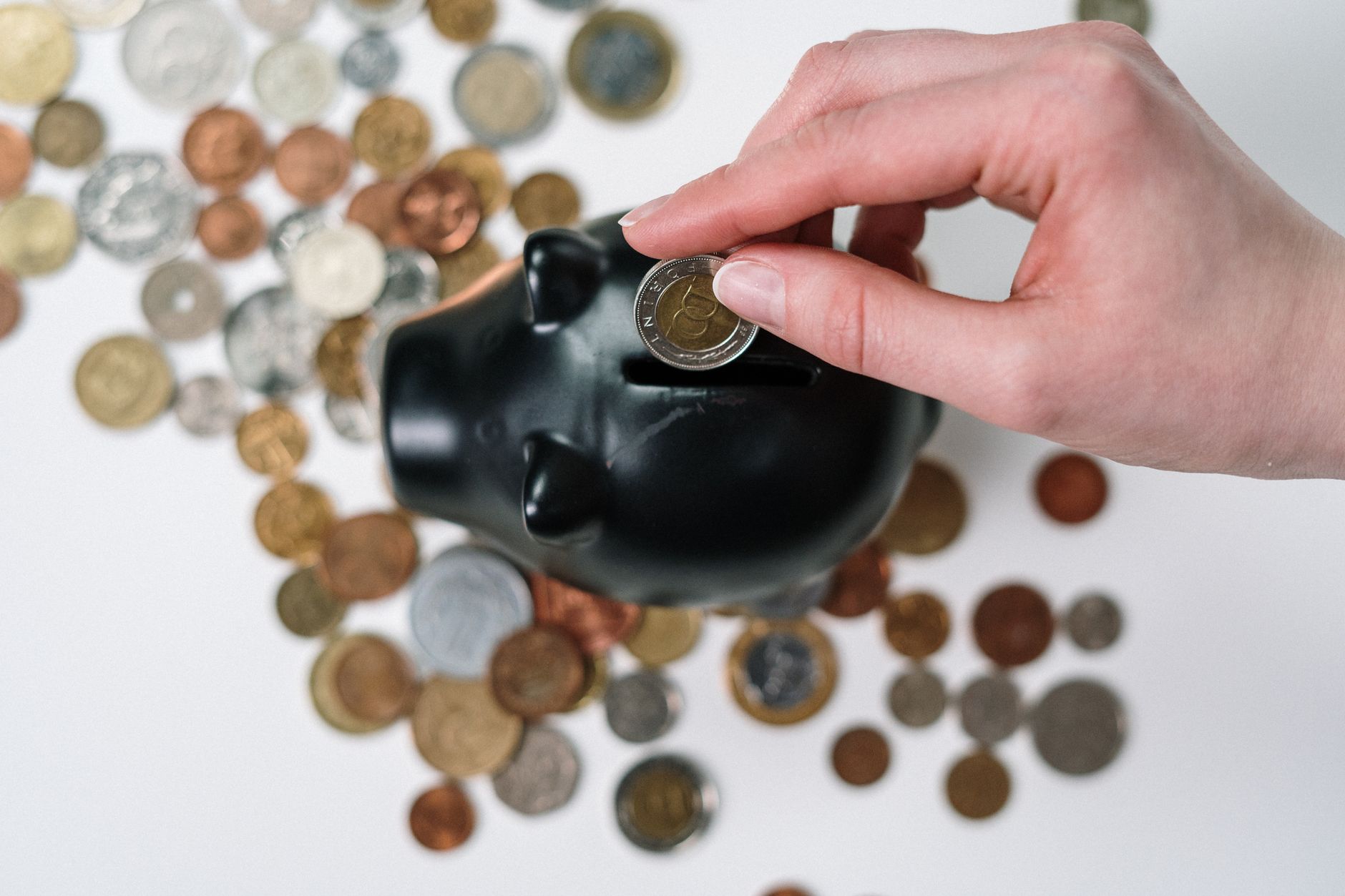 Une main tient une pièce au-dessus de la fente d’une tirelire cochon noire, entourée de pièces de monnaie variées sur un fond blanc.